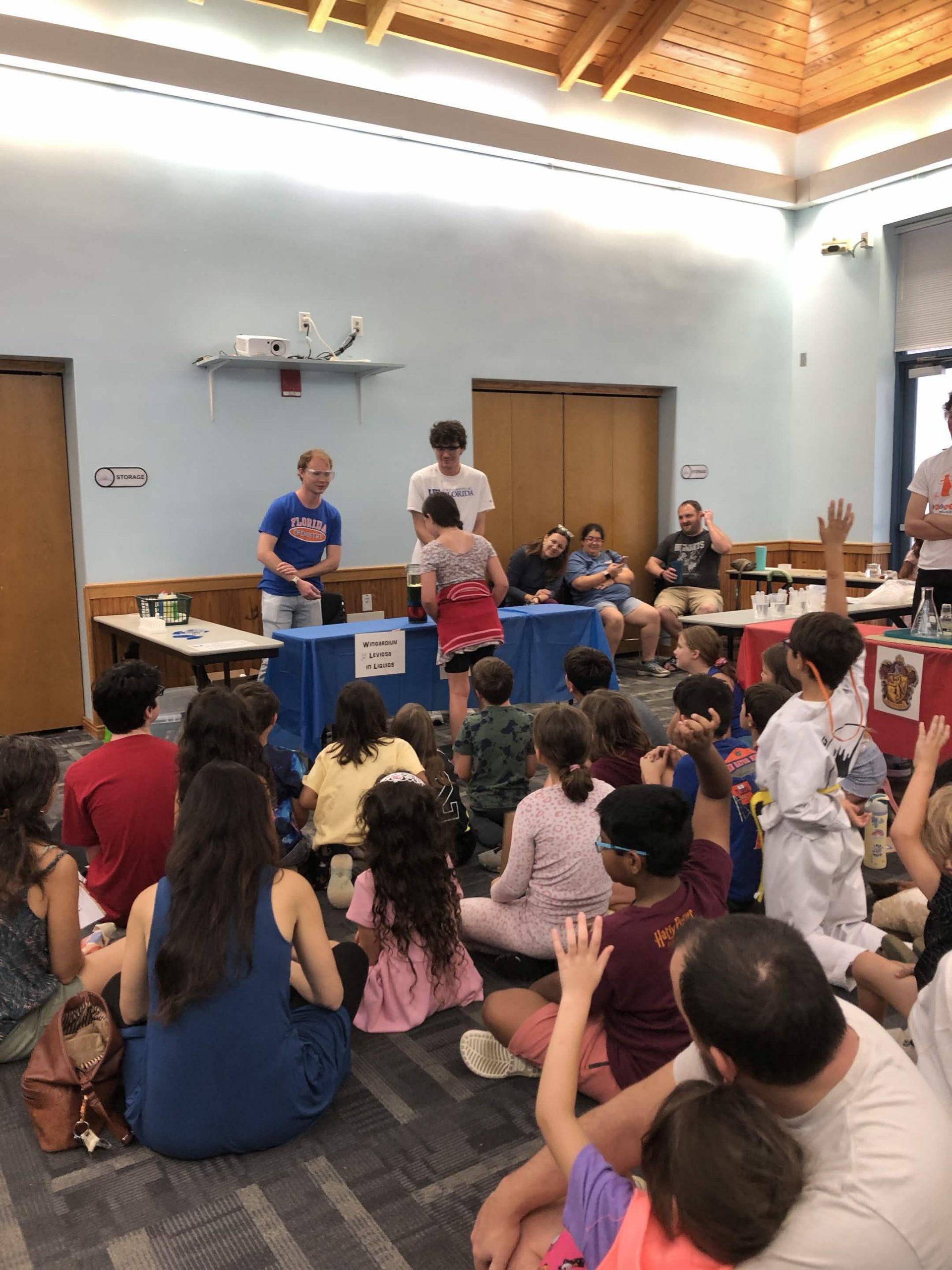 A group of young students and parents sitting on the carpet attentively watching a chemistry demonstration at a nearby table.