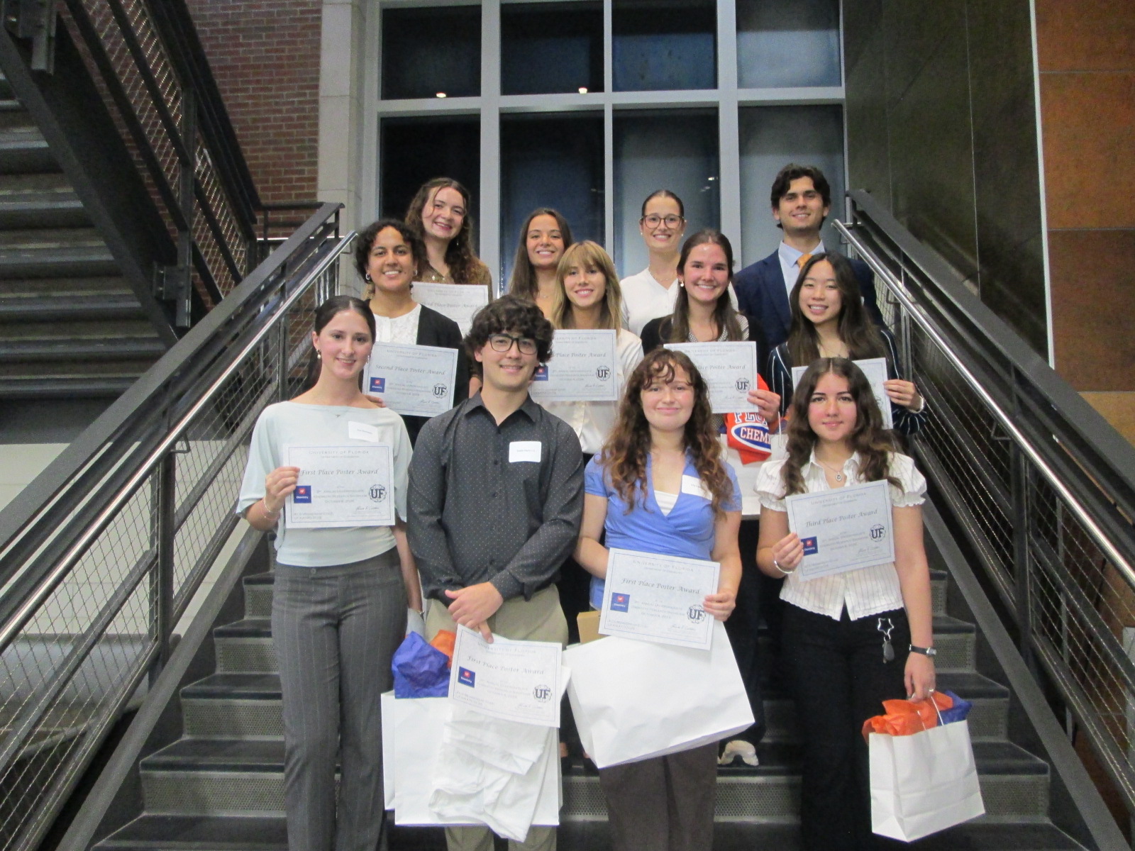 A group of 12 smiling students standing on a staircase holding award certificates and gift bags with orange and blue paper.