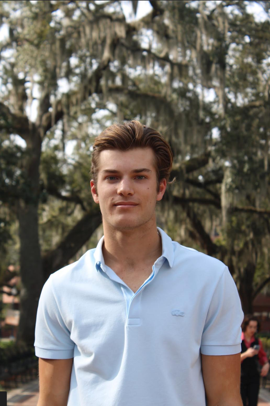 A person wearing a polo shirt with a Lacoste logo stands outdoors in front of trees draped with Spanish moss.